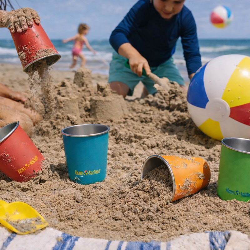Children playing on a sandy beach with colorful buckets and a ball, and steel cups.