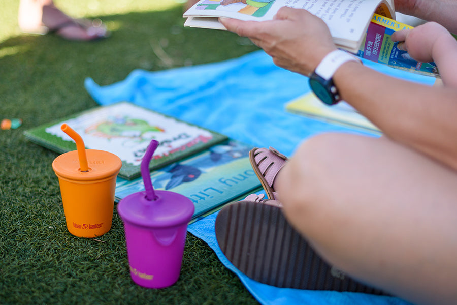 Person reading a book with colorful sippy cups on a grassy surface