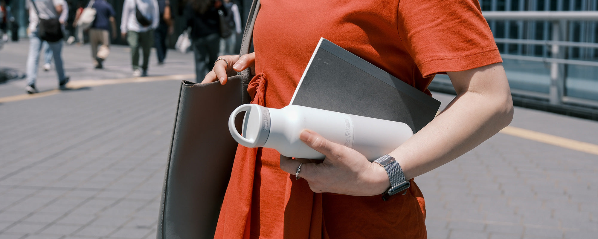 Person in orange shirt holding a water bottle and bag on a city street.
