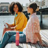 Two women sitting by a waterfront, smiling and holding water bottles