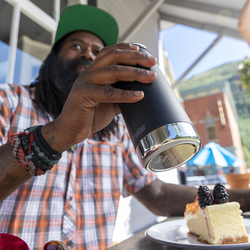 Person holding a black insulated tumbler outdoors with a slice of cake on a plate.