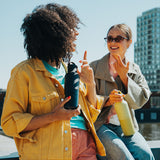 Two women sitting by a waterfront, holding water bottles, with a cityscape in the background.