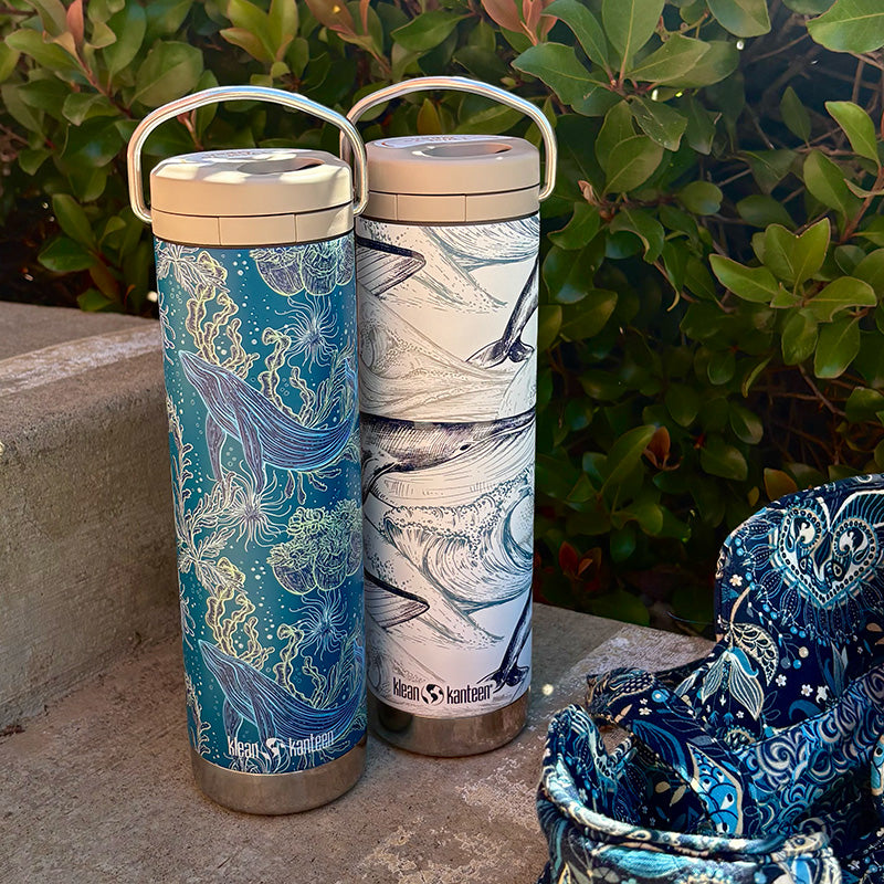 Two insulated tumblers with ocean-themed designs on a stone surface with greenery in the background.
