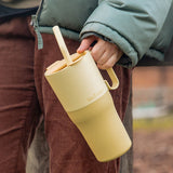 Person holding a yellow tumbler with a straw, wearing a green jacket and brown pants.