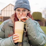 Person wearing a gray knit hat and green jacket sipping straw from a yellow tumbler.