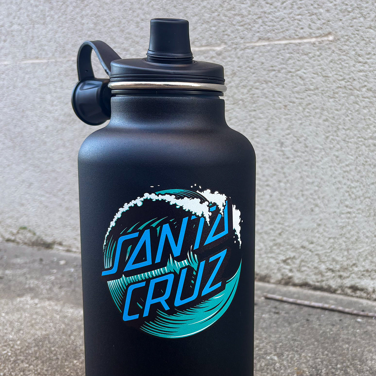 A matte black Santa Cruz water bottle with a black lid and handle, featuring a blue and teal wave graphic logo on the front, set against a light gray concrete wall.
