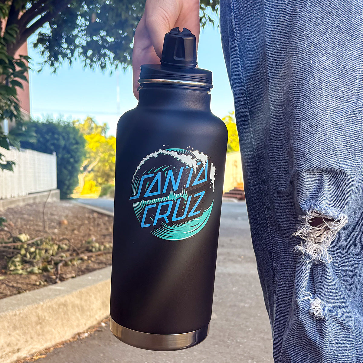 A person outdoors holding a matte black Santa Cruz water bottle with a blue and white graphic logo. The person is wearing ripped blue jeans, and trees, a fence, and a sidewalk are visible in the background.