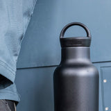 Black water bottle with loop cap held by a person wearing a light blue jacket against a gray background