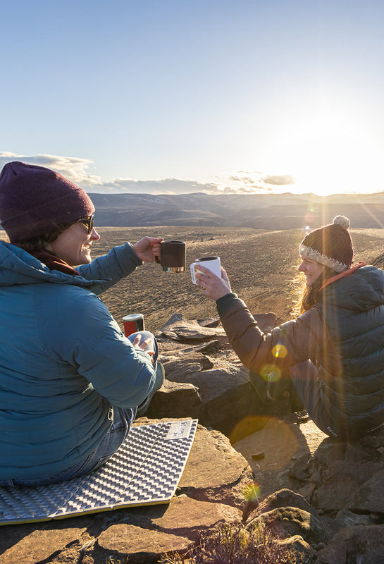Two women at sunrise camping with coffee mugs by canyon
