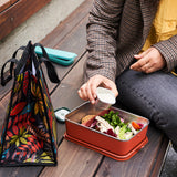 Person preparing a salad in a metal lunch box with a colorful bag on a wooden surface