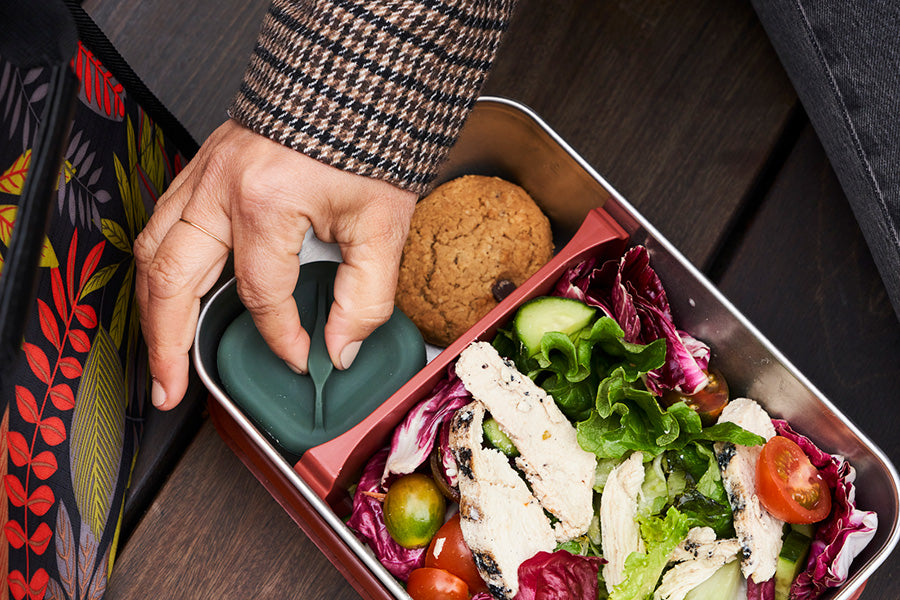 Person reaching into a lunchbox with salad and cookie on a wooden surface