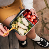 Person holding a bento box with salad and strawberries on a stone pavement.