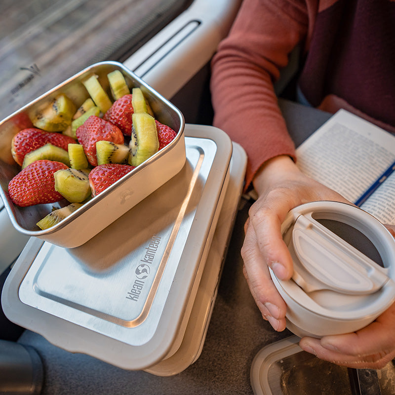 Person holding a container of fruit next to a reusable coffee cup on a car seat.