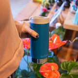 Person holding a blue insulated tumbler with plants in the background