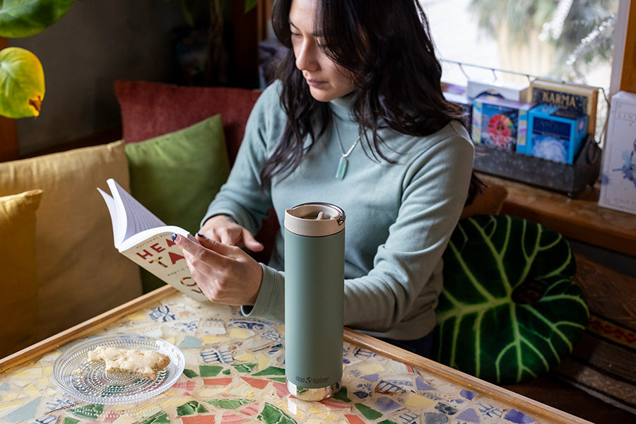 Woman reading a book with a green tumbler on a colorful table in a cozy setting.