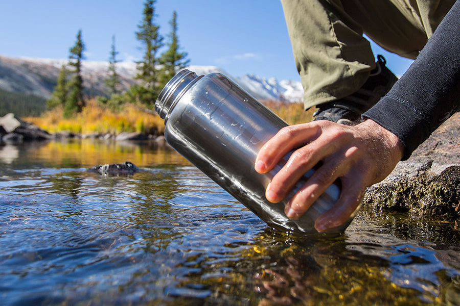 Person filling a water bottle from a stream with mountains in the background