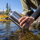 Person filling a stainless steel water bottle from a stream with mountains in the background