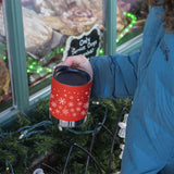 Person holding a red insulated camp mug with snowflake pattern in front of a window display.