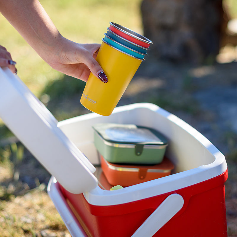 Person holding a yellow insulated mug with multiple color layers over a red cooler outdoors.