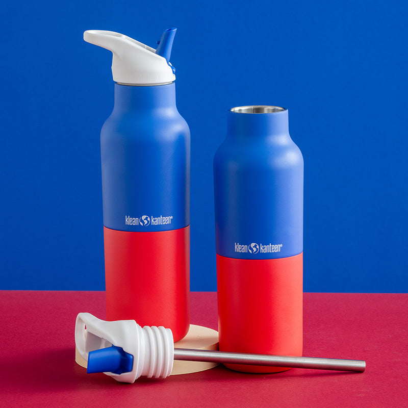 Two Klean Kanteen insulated water bottles in a blue and red color scheme, shown on a red surface against a blue background, with a white lid and steel straw placed in front.