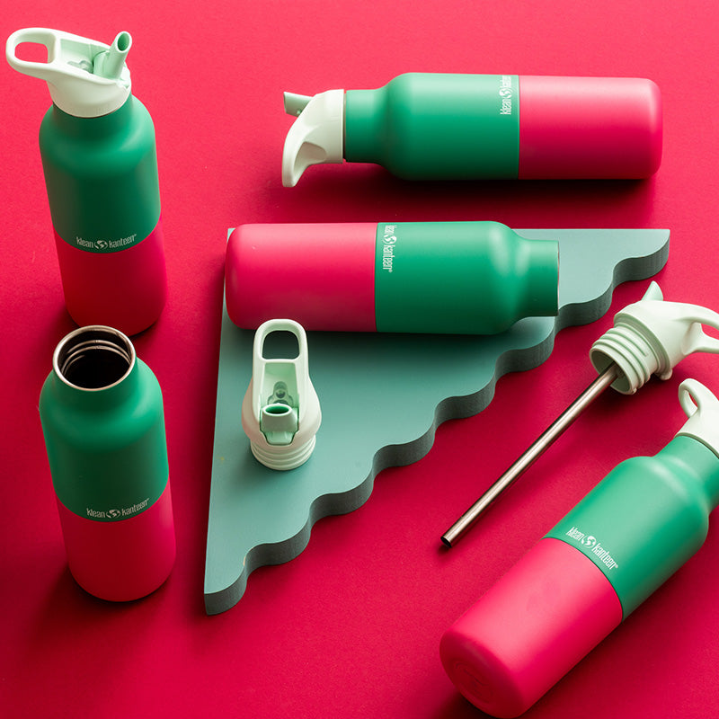 A set of insulated water bottles and bottle accessories arranged on a bright pink background. The bottles are green and pink with light green caps and lids and steel straw