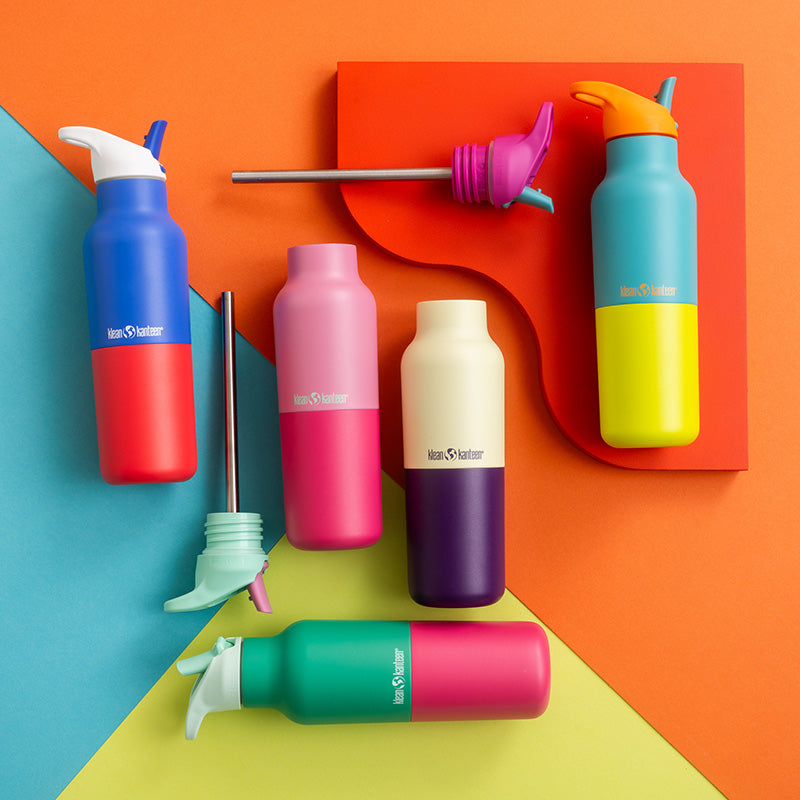 Six reusable insulated bottles and a cap with steel straw in a colorful flat-lay on a geometric orange, blue, yellow, and green background