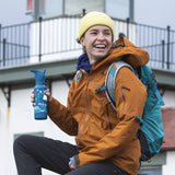 Person wearing a brown jacket and yellow beanie, holding a blue water bottle with a building in the background.