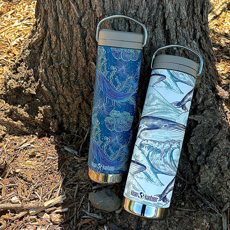 Two insulated bottles with whale designs leaning against a tree trunk.