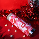 Pink and red valentine's botlte on side with heart designs on a red background with tinsel and candy hearts.