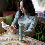 Woman reading a book with a colorful holiday tumbler on a table in a cozy setting