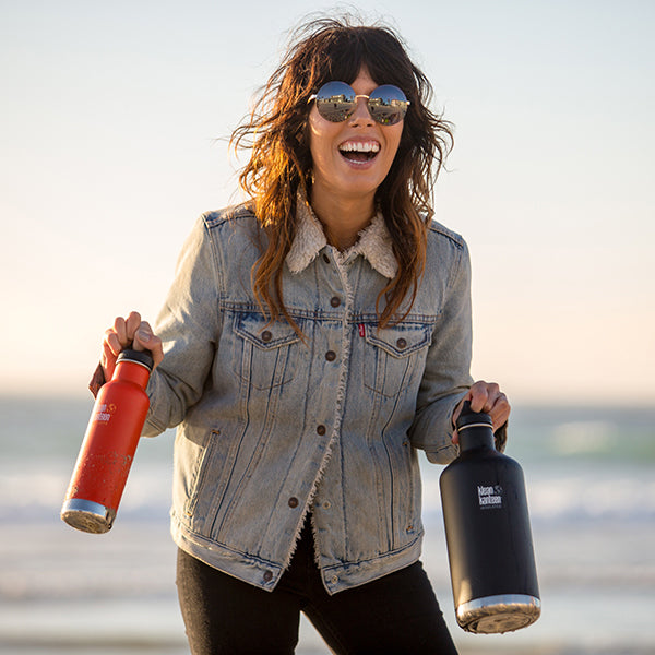 Woman on a beach holding a red water bottle and a black Klean Kanteen bottle, wearing a denim jacket.