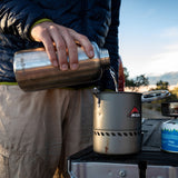 Person pouring from a water bottle into an MSR camping stove outdoors.