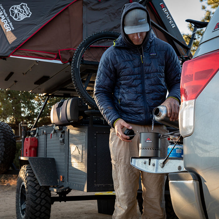 Person preparing a camping setup on a vehicle with a trailer and large water bottle