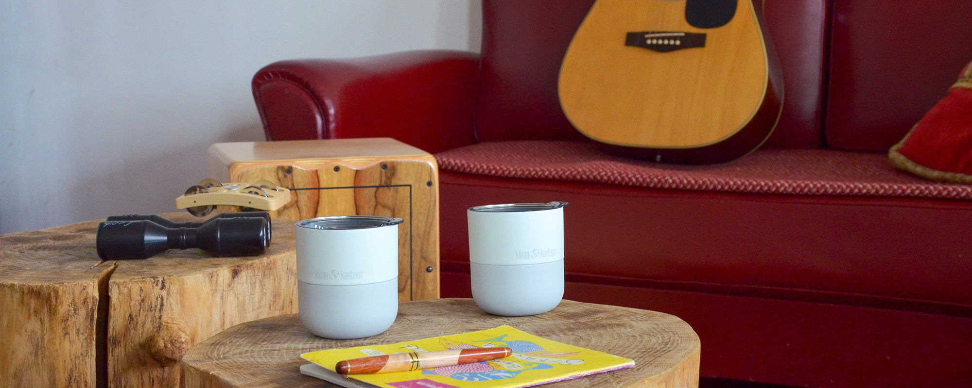 Two light blue lowball tumblers on a wooden table with a guitar leaning against a red couch.