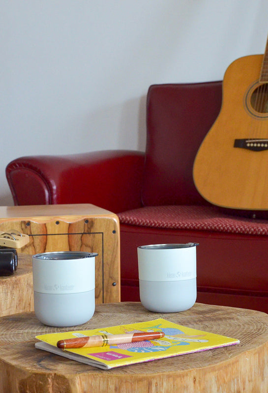 Two light blue lowball tumblers on a wooden table with a guitar leaning against a red couch.