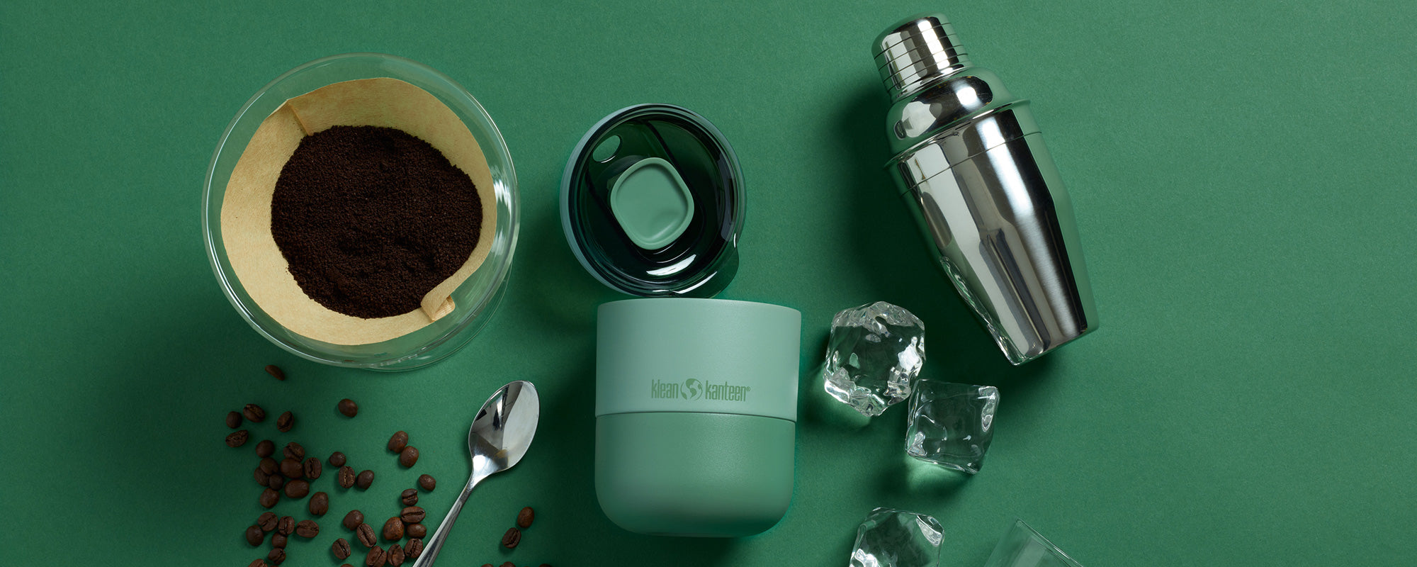 Green tumbler, coffee filter, coffee beans, spoon, and cocktail shaker on a green background
