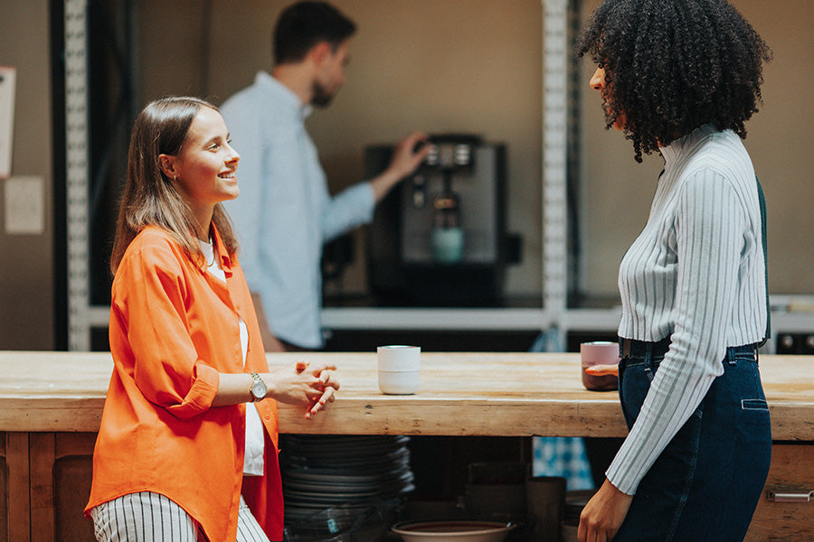 Two women engaged in a conversation at a coffee shop counter with a barista in the background, and two tumblers