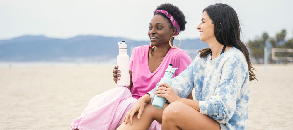 Two women on beach drinking from water bottles