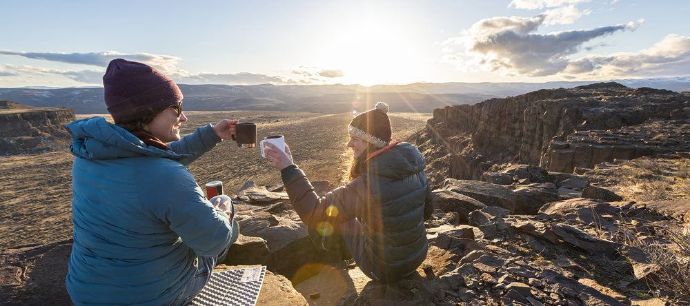 Two women at sunrise camping with coffee mugs by canyon