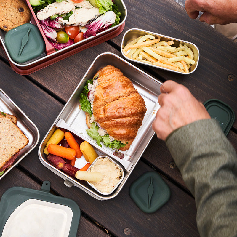Lunch containers with a croissant, salad, and other food items on a wooden table.