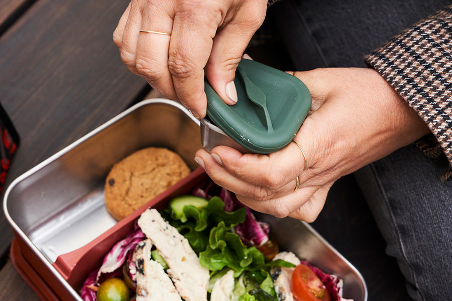 Person opening a small food box with a green lid,  with lunch box in background containing a salad and cookies.