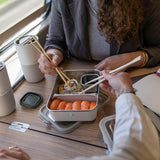 Two people enjoying sushi from a portable container with chopsticks on a wooden table.