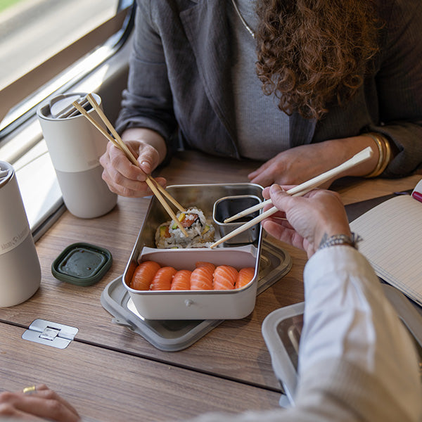 Two people enjoying sushi from a portable container with chopsticks on a wooden table.