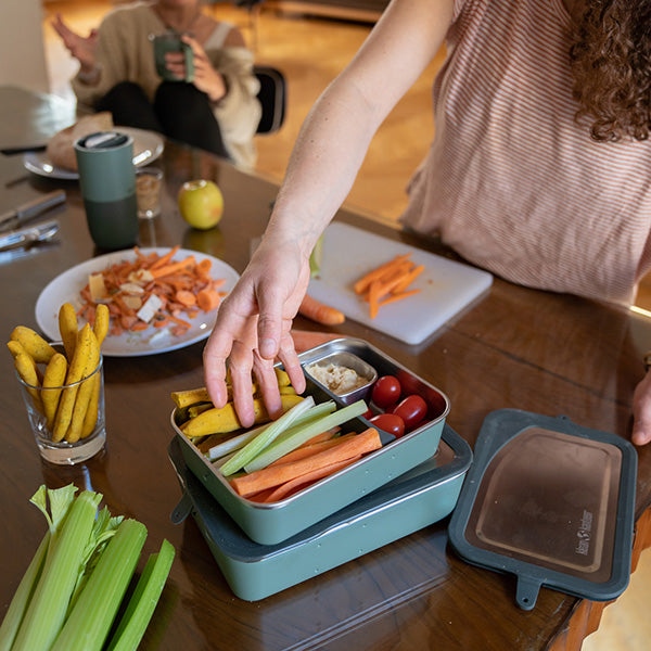 Person reaching for snacks from a bento box on a wooden table with a casual setting.