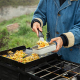 Person cooking outdoors, transferring food from a pan to a container on a grill.