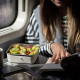 Person reading a book with a container of fruit on a table inside a vehicle.