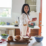 Woman in a kitchen preparing food with bread and fruits on a counter.