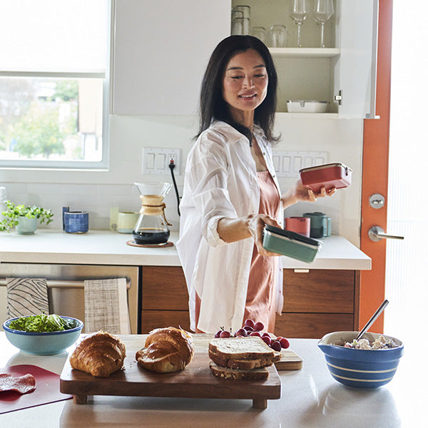 Woman in a kitchen preparing food with bread and fruits on a counter.